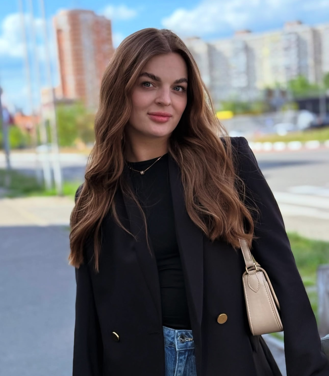 A woman with wavy, shoulder-length brown hair is smiling gently. She is wearing a white blouse and is standing in a bright, minimalist setting with turquoise decorative items in the background.